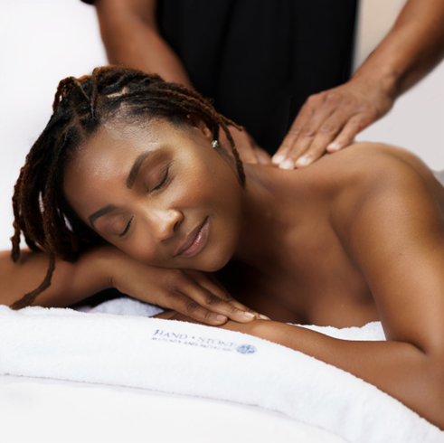 A woman with closed eyes rests her head on her hands on top of a white Hand & Stone towel as someone massages her shoulders 